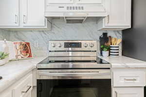 Kitchen with custom exhaust hood, electric stove, light stone countertops, and white cabinetry