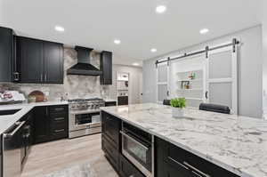 Kitchen with decorative backsplash, light hardwood / wood-style flooring, wall chimney range hood, a barn door, and stainless steel appliances