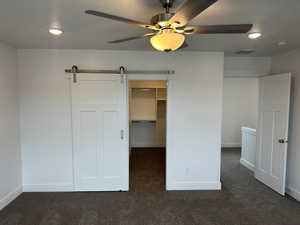 Unfurnished bedroom with a spacious closet, dark carpet, a closet, ceiling fan, and a barn door