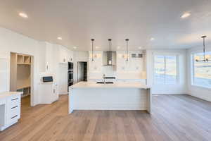 Kitchen featuring wall chimney range hood, sink, appliances with stainless steel finishes, white cabinetry, and hanging light fixtures