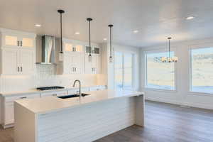 Kitchen with wall chimney range hood, sink, white cabinetry, hanging light fixtures, and a kitchen island with sink