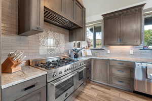 Kitchen featuring decorative backsplash, appliances with stainless steel finishes, light stone countertops, premium range hood, and light hardwood / wood-style floors