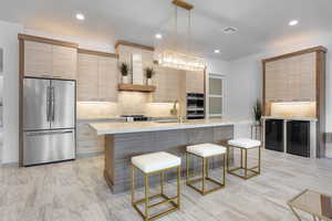 Kitchen with tasteful backsplash, an island with sink, decorative light fixtures, light brown cabinetry, and appliances with stainless steel finishes