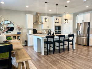 Kitchen featuring appliances with stainless steel finishes, white cabinets, wall chimney exhaust hood, an island with sink, and light wood-type flooring