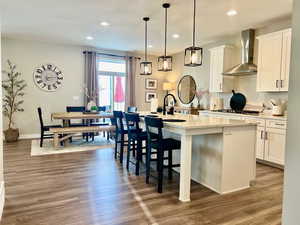 Kitchen with a kitchen island with sink, wall chimney range hood, white cabinets, hardwood / wood-style flooring, and decorative light fixtures