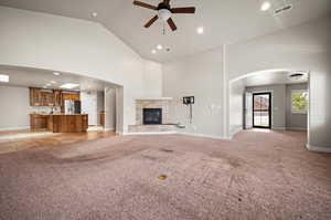 Spacious Great room featuring a tiled fireplace, high vaulted ceiling, ceiling fan, light colored carpet with electrical outlet on floor, view of front entry and kitchen