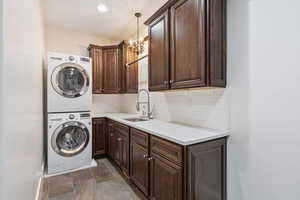 Clothes washing area with dark tile patterned floors, cabinets, an inviting chandelier, stacked washer and dryer, and sink