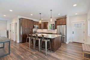 Guest house kitchen with appliances with stainless steel finishes, dark wood-type flooring, hanging light fixtures, a center island, and sink