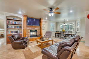 Living room with light tile patterned flooring, ceiling fan with notable chandelier, a stone fireplace, and crown molding