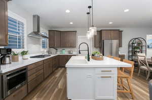 Kitchen featuring pendant lighting, appliances with stainless steel finishes, a kitchen island with sink, dark hardwood / wood-style floors, and wall chimney range hood