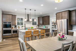 Kitchen with wall chimney range hood, pendant lighting, a center island with sink, stainless steel appliances, and wood-type flooring