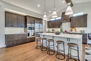 Kitchen with custom range hood, stainless steel appliances, decorative backsplash, dark brown cabinetry, and light wood-type flooring