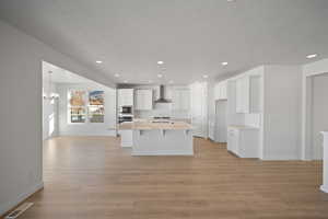 Kitchen with a center island with sink, wall chimney range hood, sink, light wood-type flooring, and white cabinetry