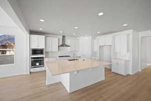 Kitchen featuring wall chimney exhaust hood, stainless steel appliances, sink, a center island with sink, and white cabinetry