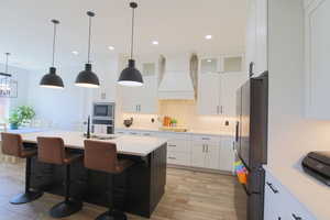 Kitchen with white cabinets, premium range hood, a center island with sink, light hardwood / wood-style flooring, and appliances with stainless steel finishes