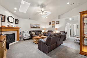 Living room with carpet floors, a tiled fireplace, vaulted ceiling with skylight, and ceiling fan