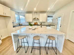 Kitchen featuring white cabinetry, wall chimney range hood, light hardwood / wood-style flooring, an island with sink, and a breakfast bar