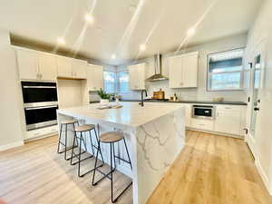 Kitchen featuring light wood-type flooring, wall chimney exhaust hood, stainless steel appliances, white cabinets, and an island with sink