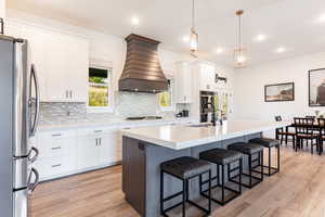 Kitchen with light wood-type flooring, white cabinetry, appliances with stainless steel finishes, and custom range hood