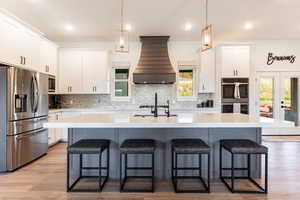 Kitchen featuring appliances with stainless steel finishes, light hardwood / wood-style flooring, custom range hood, decorative backsplash, and white cabinets