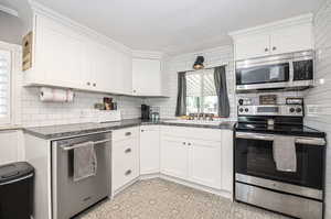 Kitchen featuring farmhouse sink, white cabinets with crown molding, tasteful backsplash, and stainless steel appliances.