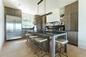 Kitchen featuring backsplash, light wood-type flooring, stainless steel built in refrigerator, dark stone counters, and a kitchen breakfast bar