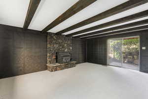 Unfurnished living room with carpet floors, beam ceiling, a stone fireplace, and a wood stove