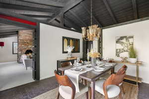 Dining space with carpet, a stone fireplace, an inviting chandelier, lofted ceiling with beams, and brick wall