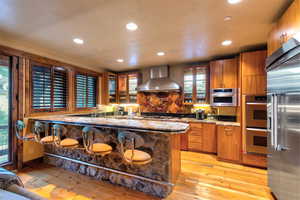 Kitchen featuring a kitchen bar, dark stone countertops, wall chimney exhaust hood, light hardwood / wood-style floors, and stainless steel appliances