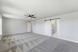 Unfurnished bedroom with a barn door, ceiling fan, light carpet, and ensuite bath