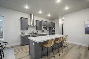 Kitchen with a kitchen island with sink, a kitchen breakfast bar, appliances with stainless steel finishes, gray cabinetry, and wall chimney range hood