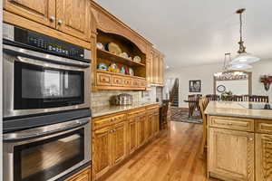 Kitchen with light wood-type flooring, stainless steel double oven, light stone countertops, tasteful backsplash, and hanging light fixtures