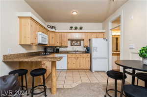 Kitchen with light brown cabinets, dark stone counters, light tile patterned floors, sink, and white appliances