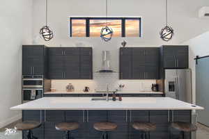 Kitchen featuring appliances with stainless steel finishes, decorative light fixtures, wall chimney range hood, and an island with sink