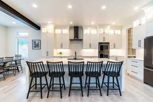 Kitchen with appliances with stainless steel finishes, light hardwood / wood-style floors, white cabinetry, wall chimney exhaust hood, and a center island with sink