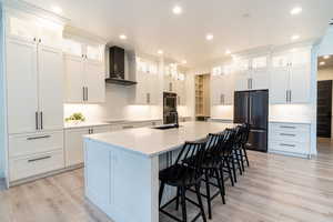 Kitchen with light hardwood / wood-style flooring, wall chimney range hood, an island with sink, and black appliances