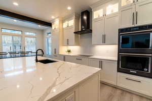 Kitchen featuring wall chimney range hood, light hardwood / wood-style flooring, light stone countertops, sink, and black appliances