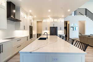 Kitchen featuring sink, light hardwood / wood-style floors, a large island with sink, wall chimney range hood, and black appliances