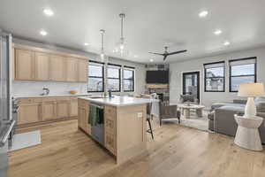 Kitchen featuring a fireplace, light wood-type flooring, an island with sink, light brown cabinetry, and stainless steel dishwasher