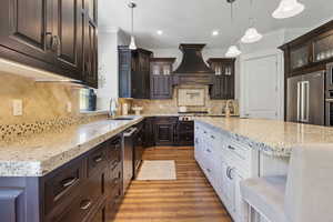 Kitchen featuring backsplash, dark brown cabinetry, custom range hood, appliances with stainless steel finishes, and light hardwood / wood-style floors