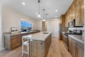 Kitchen with light wood-type flooring, decorative light fixtures, stainless steel appliances, sink, and a center island with sink