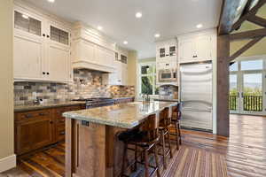 Kitchen with built in appliances, a kitchen island with sink, tasteful backsplash, dark wood-type flooring, and light stone counters