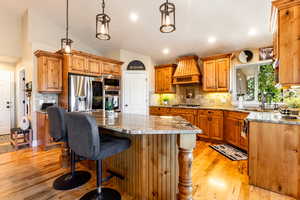 Kitchen with custom range hood, vaulted ceiling, appliances with stainless steel finishes, light stone countertops, and light hardwood / wood-style floors