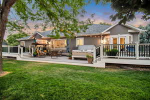 Back house at dusk with a covered hot tub, a lawn, and a wooden deck