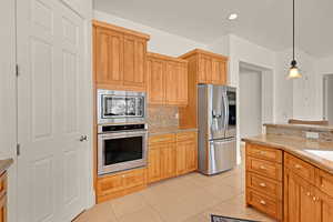 Kitchen featuring light tile patterned floors, hanging light fixtures, appliances with stainless steel finishes, light stone countertops, and tasteful backsplash