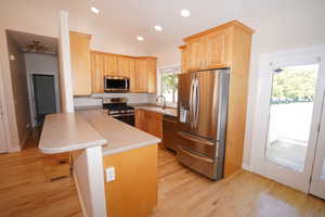 Kitchen with light hardwood / wood-style floors, vaulted ceiling, a breakfast bar, kitchen peninsula, and stainless steel appliances