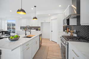 Kitchen featuring white cabinets, wall chimney range hood, hanging light fixtures, sink, and appliances with stainless steel finishes