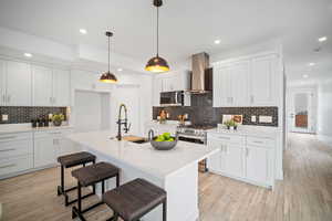 Kitchen featuring wall chimney exhaust hood, stainless steel appliances, a kitchen island with sink, sink, and white cabinetry