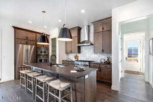 Kitchen featuring wall chimney exhaust hood, a center island with sink, hanging light fixtures, stainless steel refrigerator, and sink