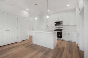 Kitchen featuring white cabinets, light hardwood / wood-style flooring, decorative light fixtures, appliances with stainless steel finishes, and a center island with sink